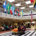 A variety of world flags hang in an atrium where community members mingle and sit at tables