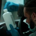 Through a van window, a man with braided hair examines a bottle of rubbing alcohol while wearing gloves to protect from COVID-19 germs