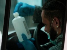 Through a van window, a man with braided hair examines a bottle of rubbing alcohol while wearing gloves to protect from COVID-19 germs