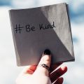 Hand holding a square piece of paper with the words written, #BeKind